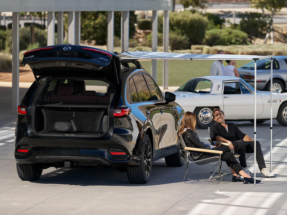 Two people sit on chairs next to a parked CX-90 with its trunk door open.