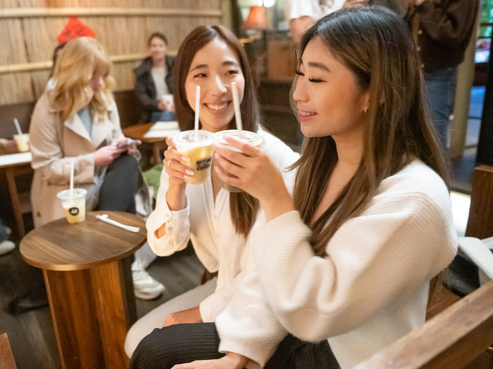 Two women hold up cups of coffee and smile while more people sit in the background. 