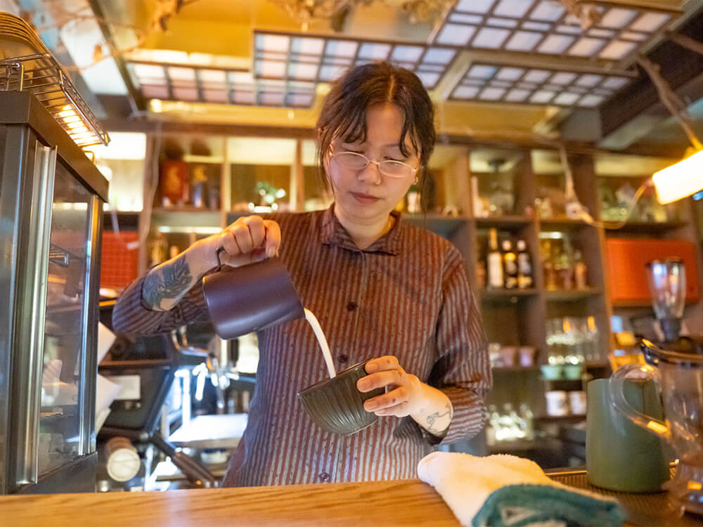 A person stands behind a coffee shop counter and pours milk into a cup of coffee. 