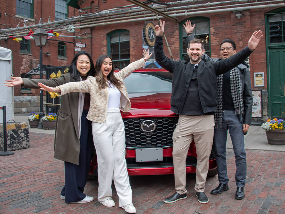 A group of people stand waving in front of a red Mazda parked outside of a coffee shop.