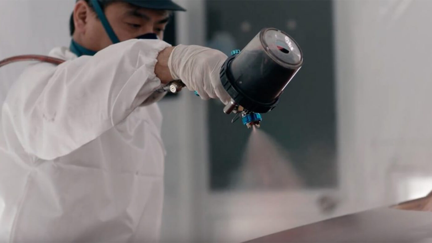 Mazda technician wearing mask and white coveralls spray paints a car part.