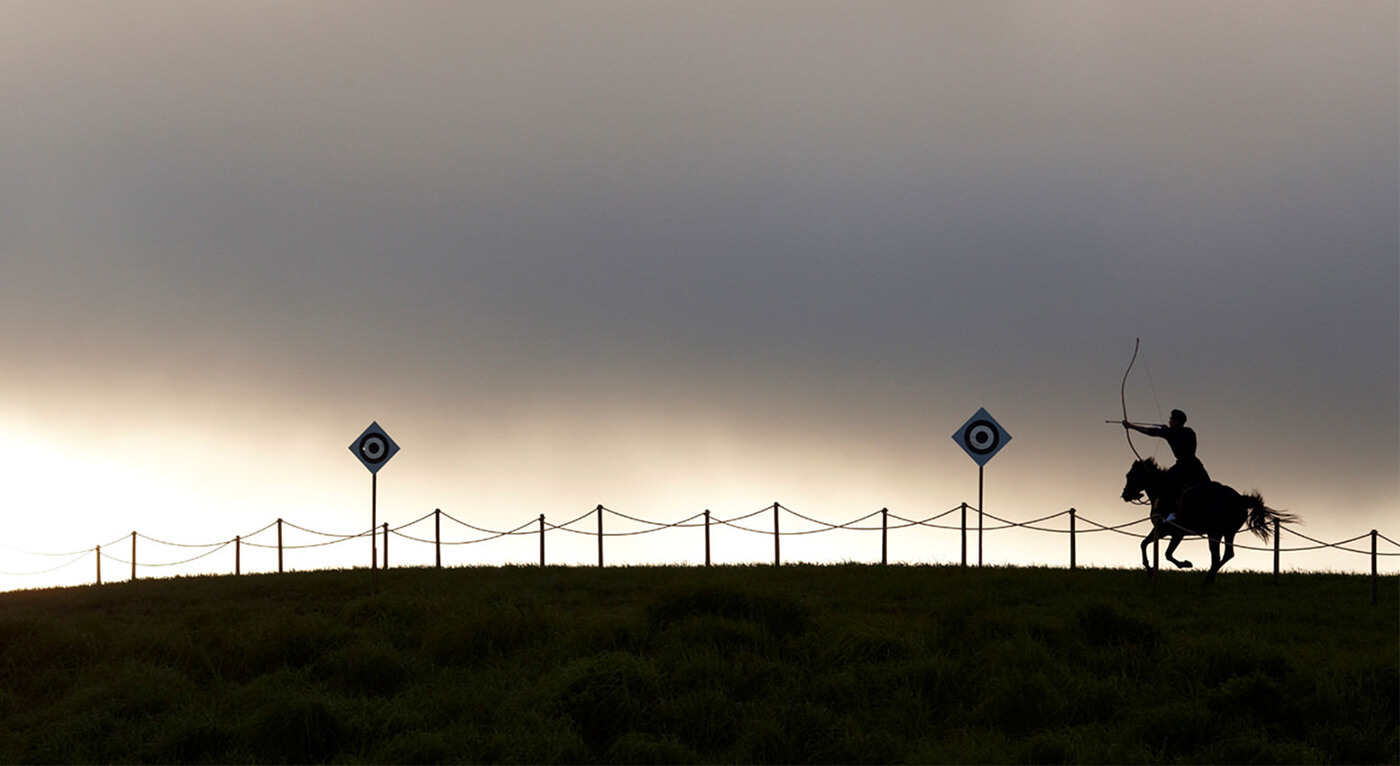 Panoramic silhouette of archer on horse moving alongside roped fencing toward and aiming for one of two bullseye targets on diamond-shaped signs. 