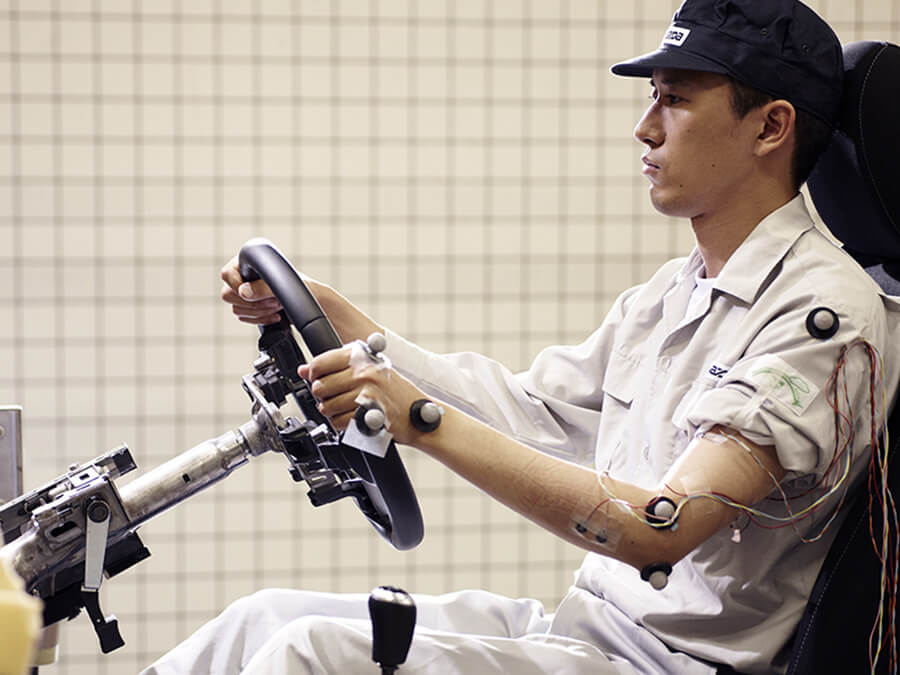 Mazda safety technician in cap and white coveralls sitting in open cockpit holding steering wheel with left sleeve rolled up and sensors taped to left hand and arm.