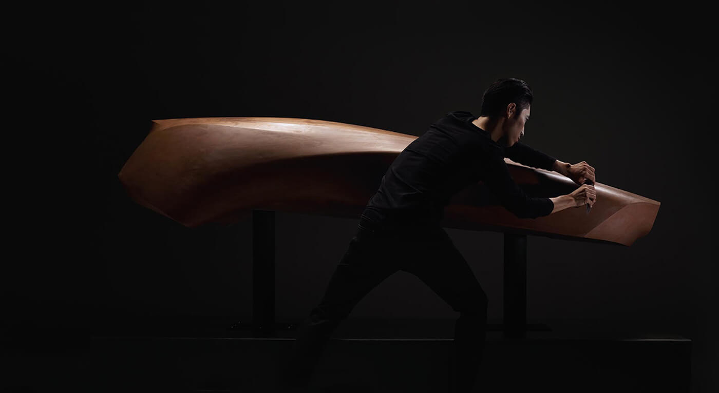 Mazda artisan dressed in black holding tool in both hands shapes a large clay car form. 