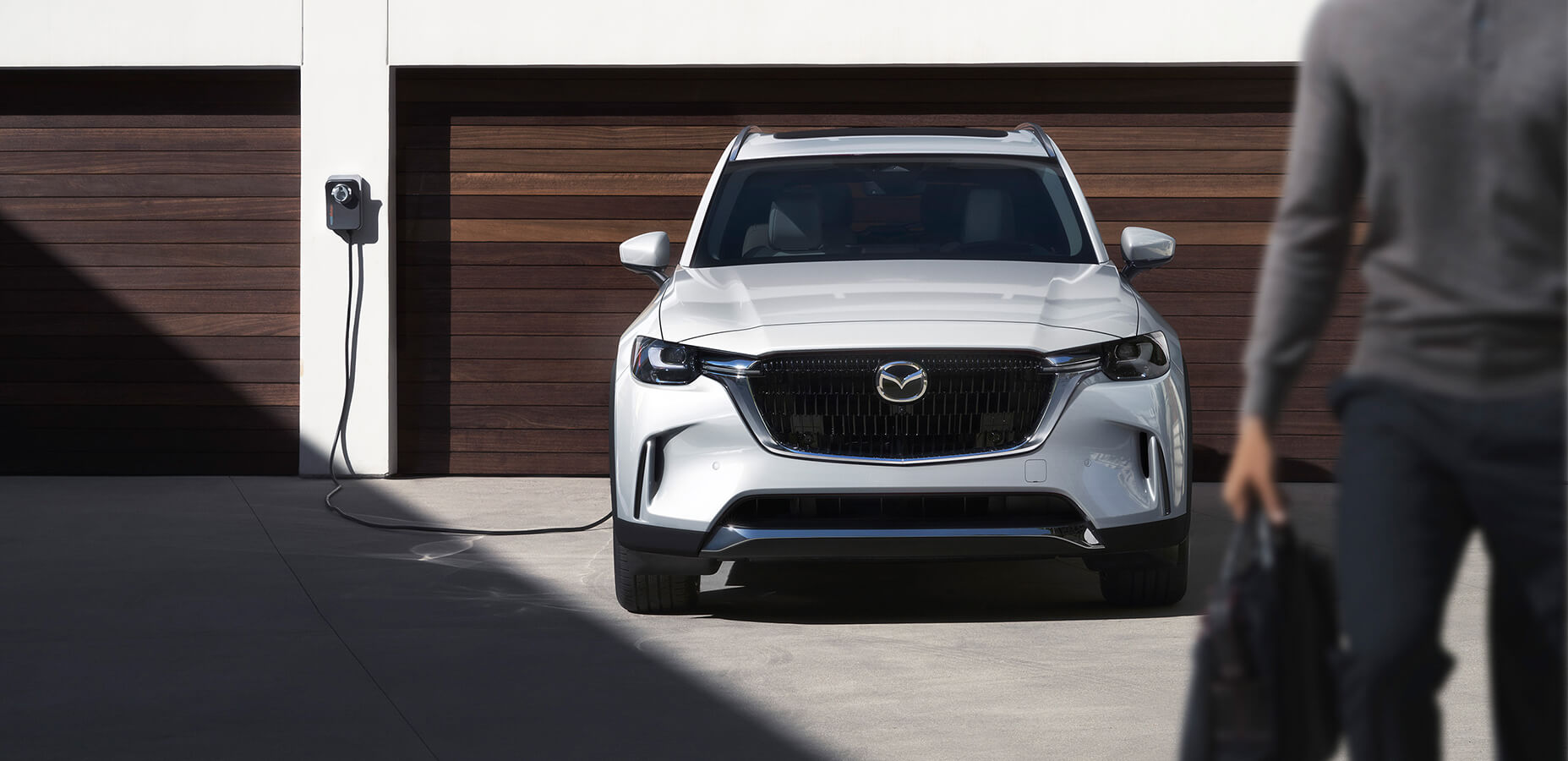 Person with tote bag walks away from CX-90 PHEV, plugged in and charging infront of two–door garage.