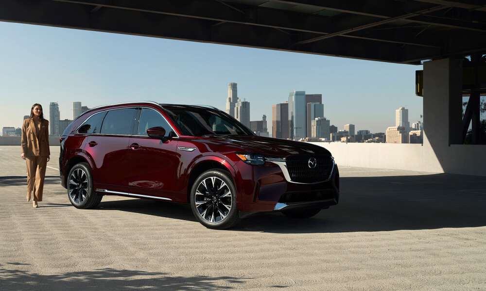 Woman in tan suit walks toward passenger side door of the CX-90 MHEV, stopped on the top floor of a parking garage. 