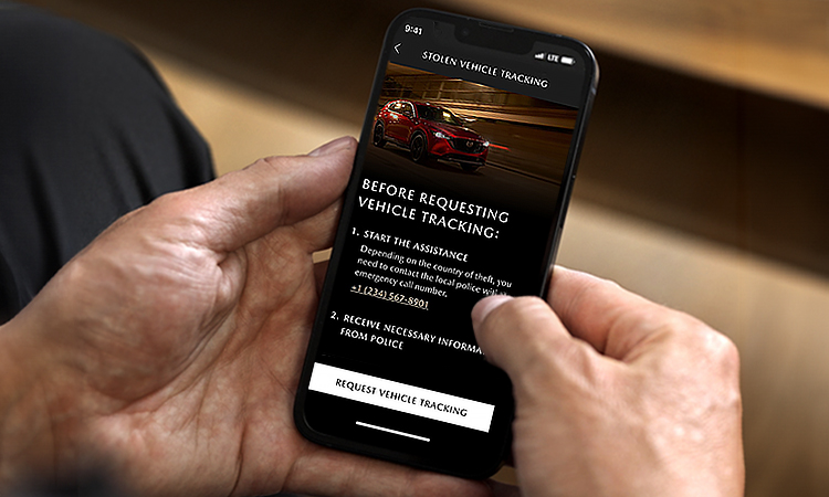 Hands holding smartphone with Mazda Stolen Vehicle Tracking on screen.