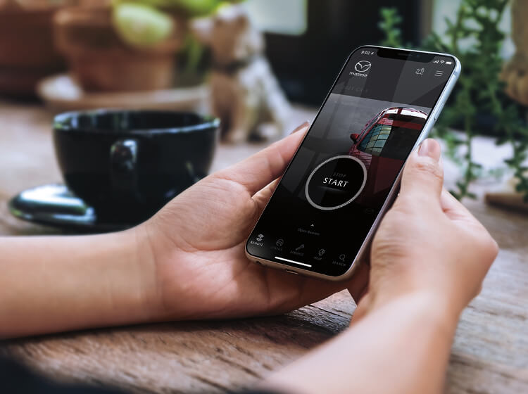 Mazda owner’s hands cradle smartphone displaying MyMazda app start screen next to black cup and saucer resting on wooden surface.
