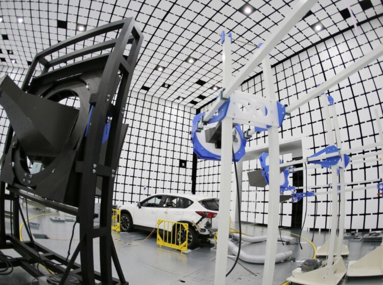 White Mazda3 with white hose attachments surrounded by equipment in a giant white room with black squares on walls 