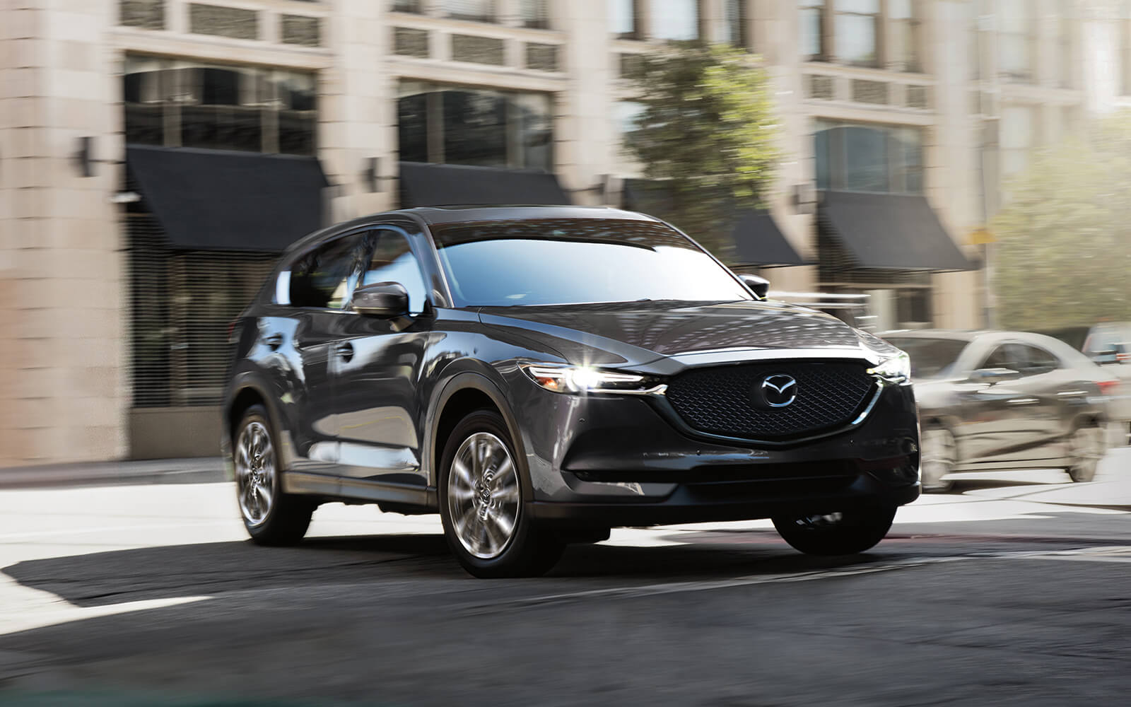 Grey Mazda SUV with headlights on turning from sunny urban street into shaded street 