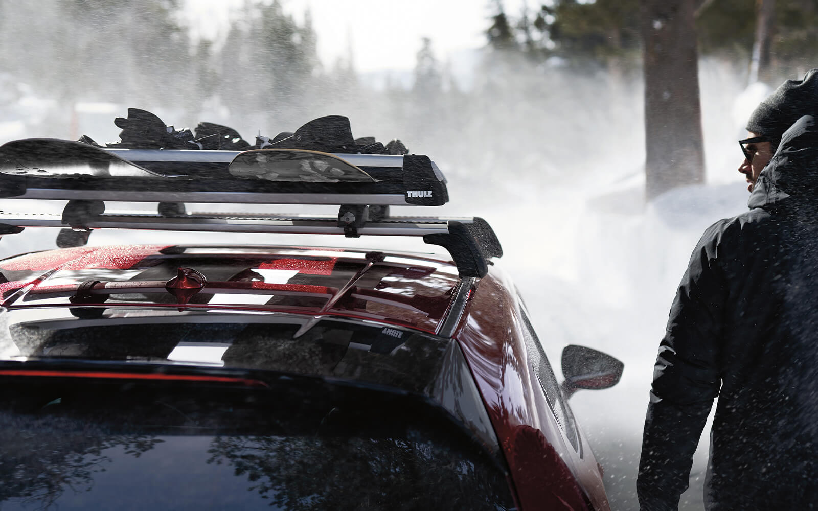 Person in winter parka walking alongside red Mazda with snowboards on roof rack and snowy landscape in distance. 