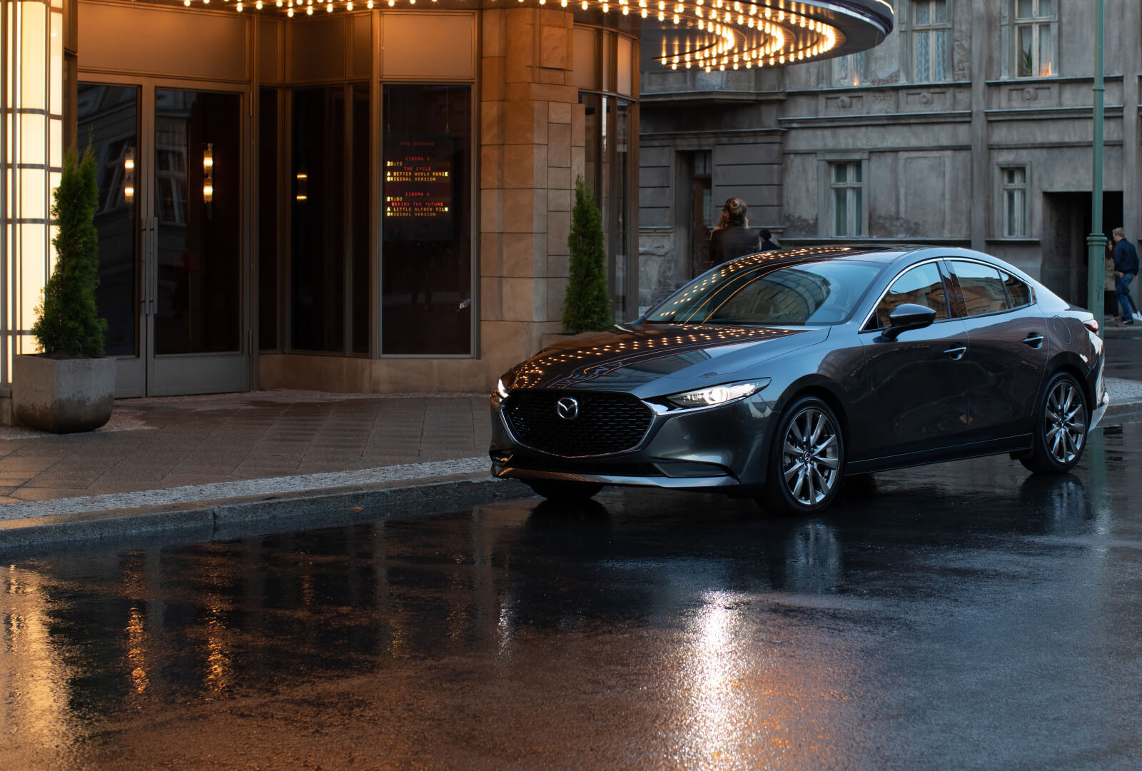 Blue Mazda3 sedan with headlights on parked on wet urban street in front of hotel.