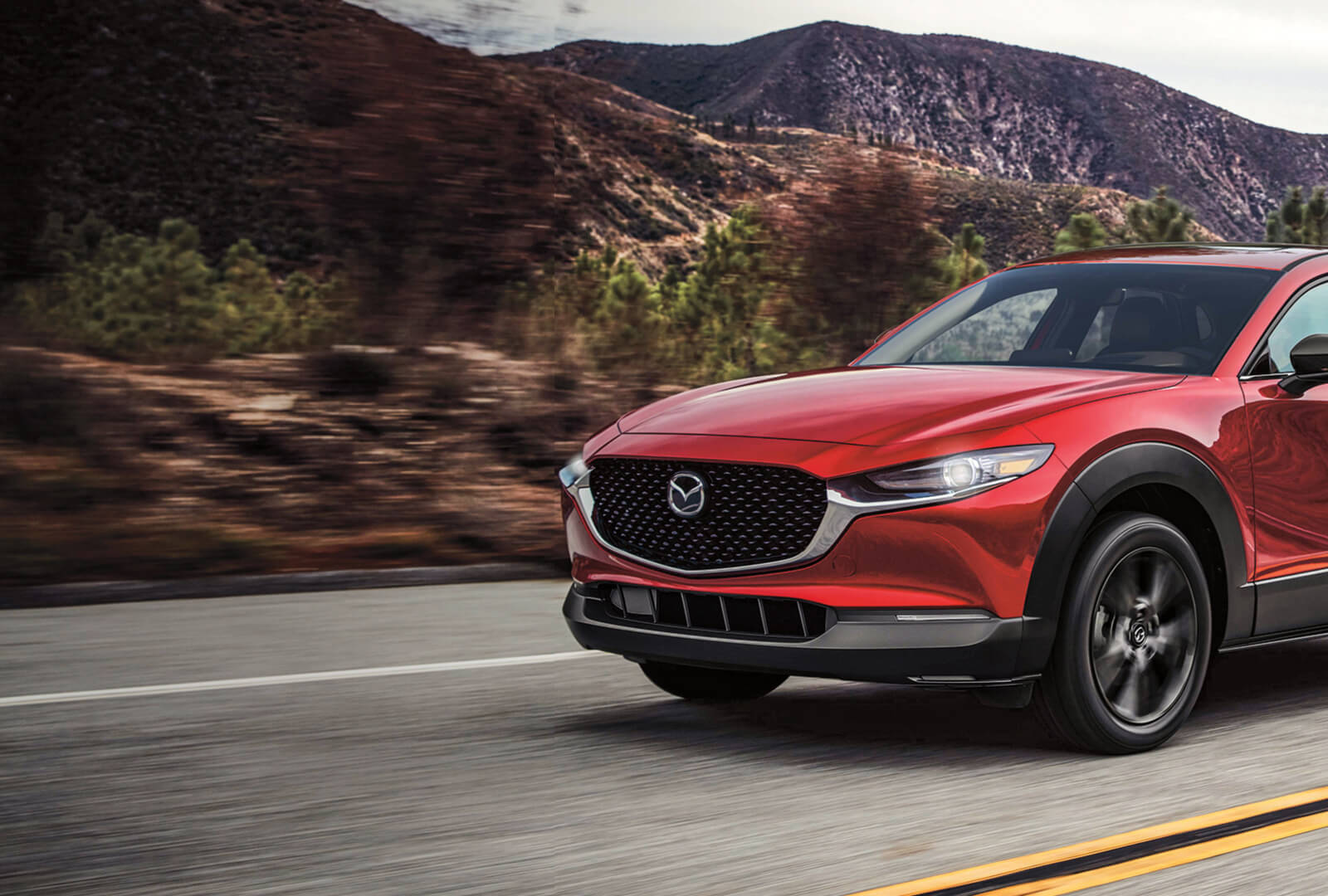 Soul red Mazda CX-5 front on highway with blurred hill country scenery in background.