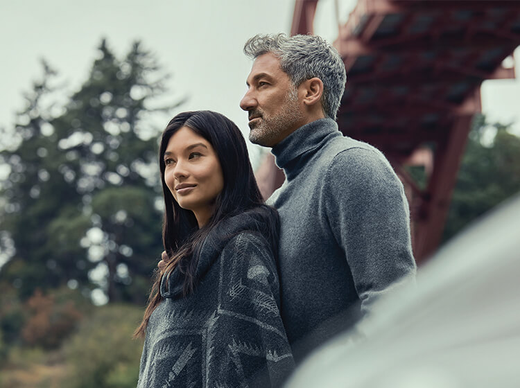 Interracial female-male couple in profile under  footbridge between blurred Mazda hood in foreground and blurred pine trees in background.