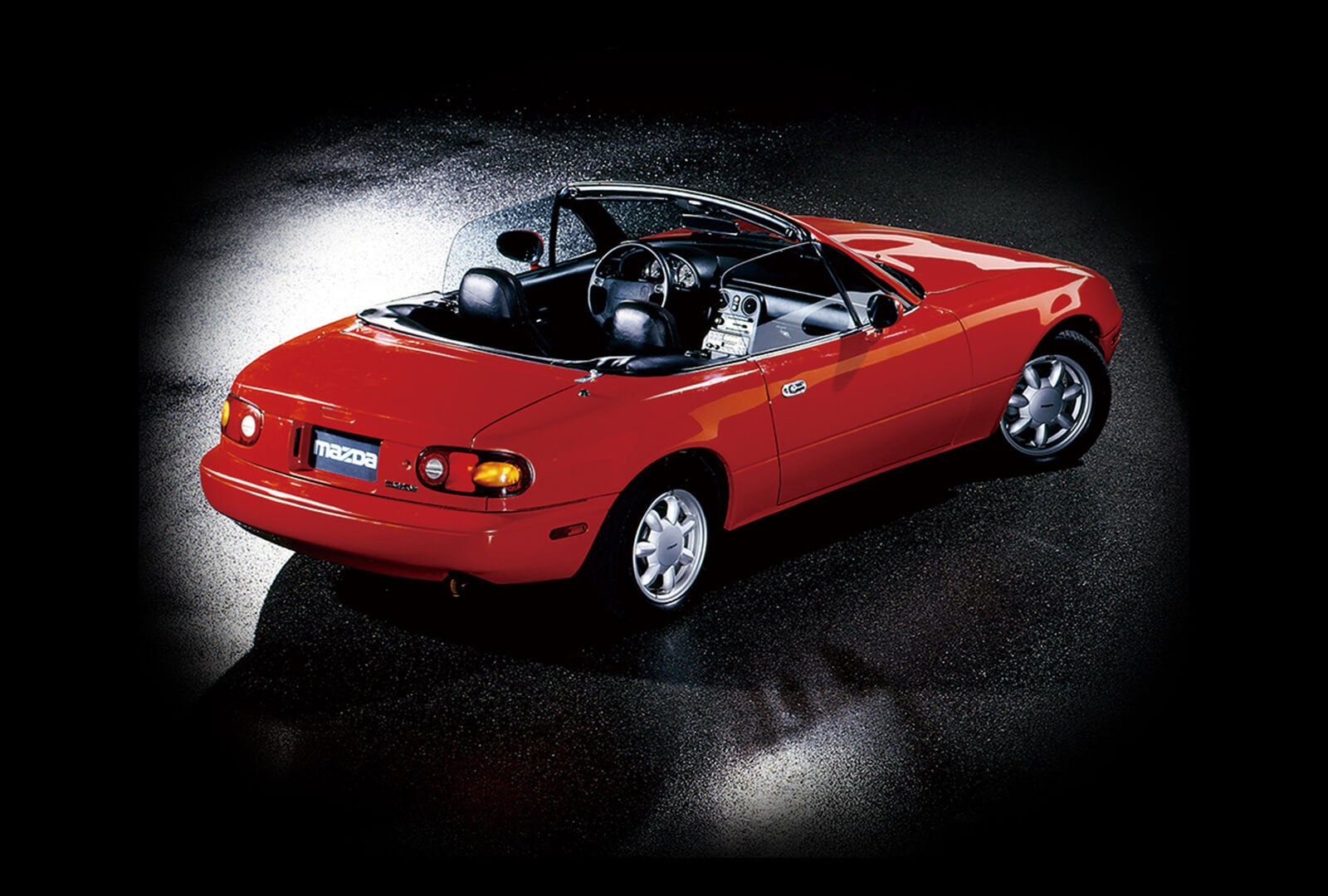 Red-orange MX-5 Miata on wet asphalt viewed from behind and slightly above passenger rear brake-light.