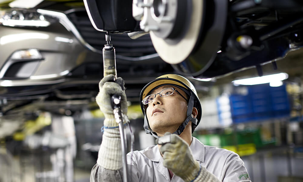 Mazda technician wearing yellow helmet and white coveralls drills into the underside of a raised Mazda vehicle.