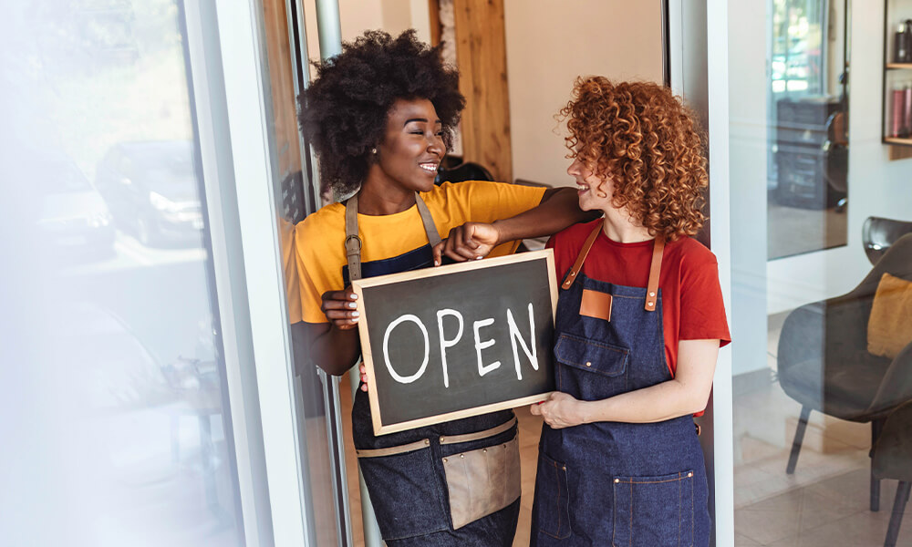 Two women in work aprons standing in the doorway to their business and proudly hold a sign saying “OPEN”