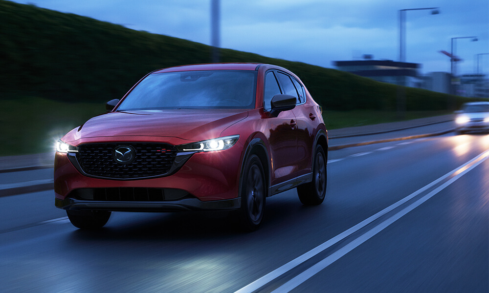 Red Mazda3 Sport with headlights on approaching on highway at twilight 