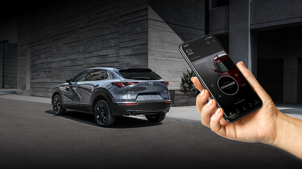 Une main tenant un téléphone sur lequel l'appli MyMazda est ouverte; un CX-30 est présent en arrière-plan