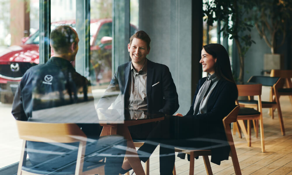 Man and woman seen through dealership window seated and smiling as they talk to a Mazda representative