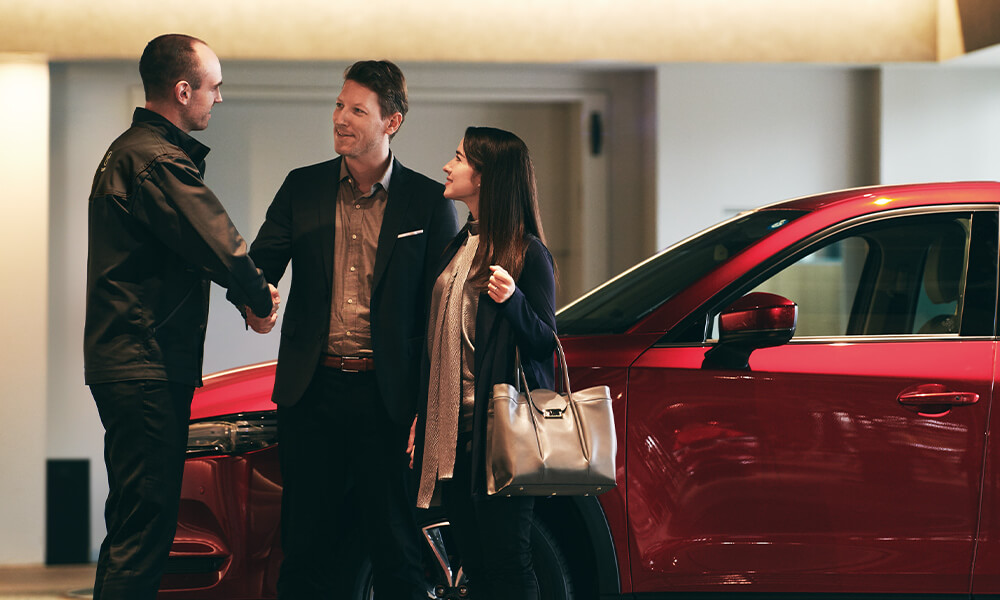 Smiling well-dressed couple shake Mazda representative’s hand  while standing in front of red Mazda vehicle.