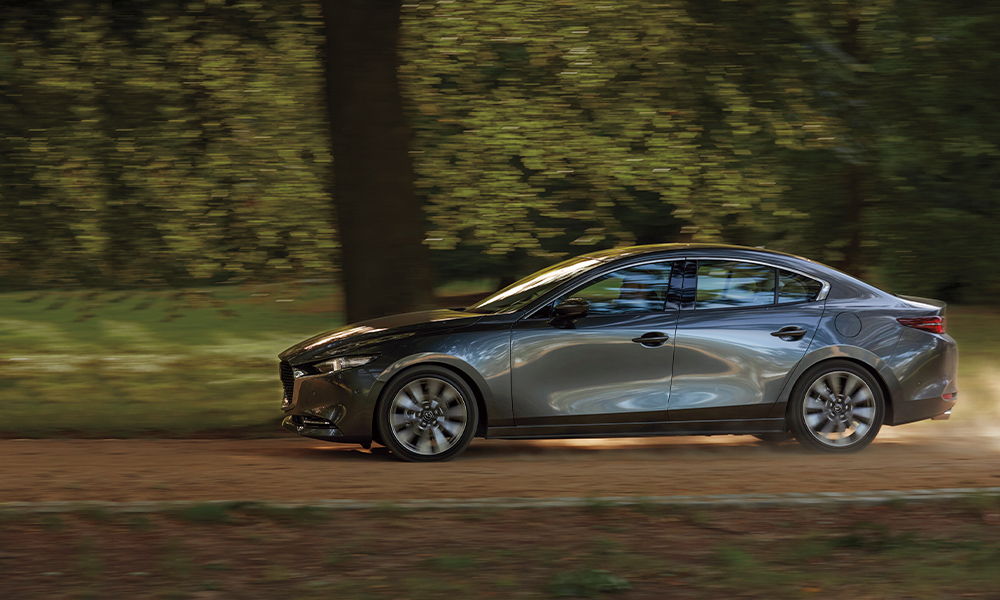 Machine grey metallic Mazda6 seen in driver-side profile driving on country road past blurred greenery in background.