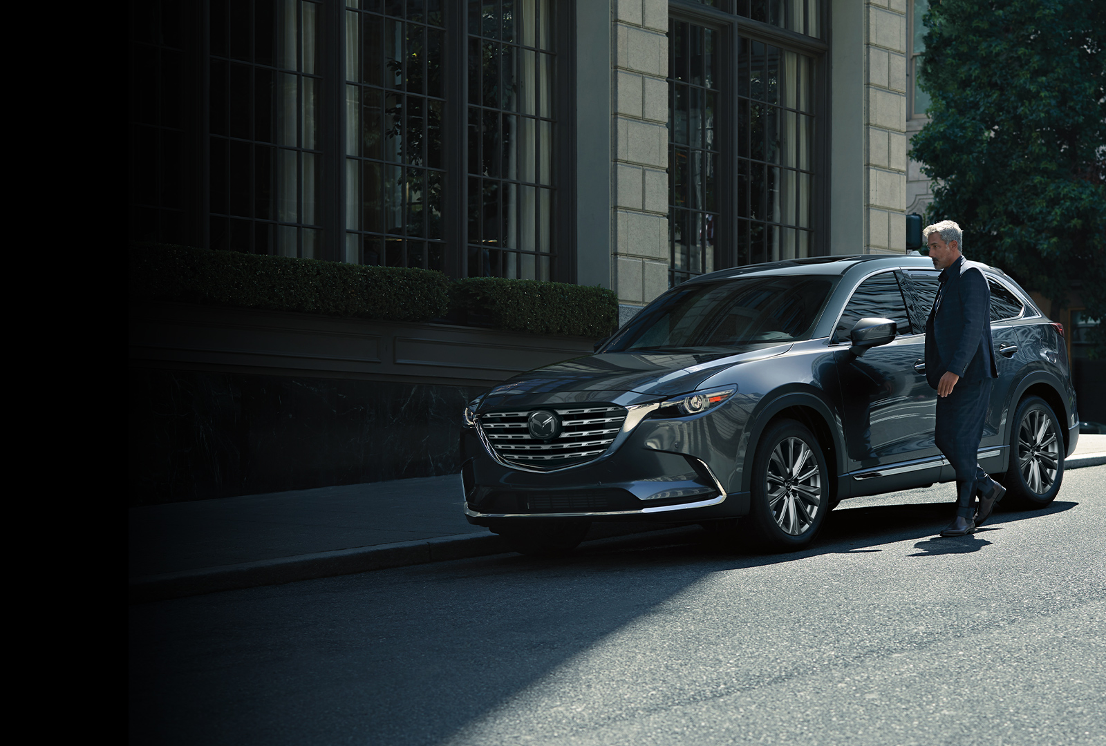 Male professional in blue suit approaching deep crystal blue mica Mazda CX-9 parked in front of building façade.