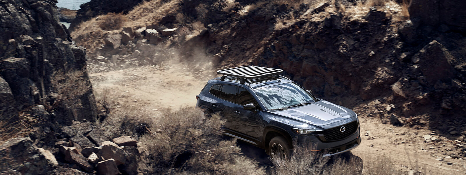 Overhead view of Mazda CX-50 with roof rack  trailing dust on dirt track between brush and rock.