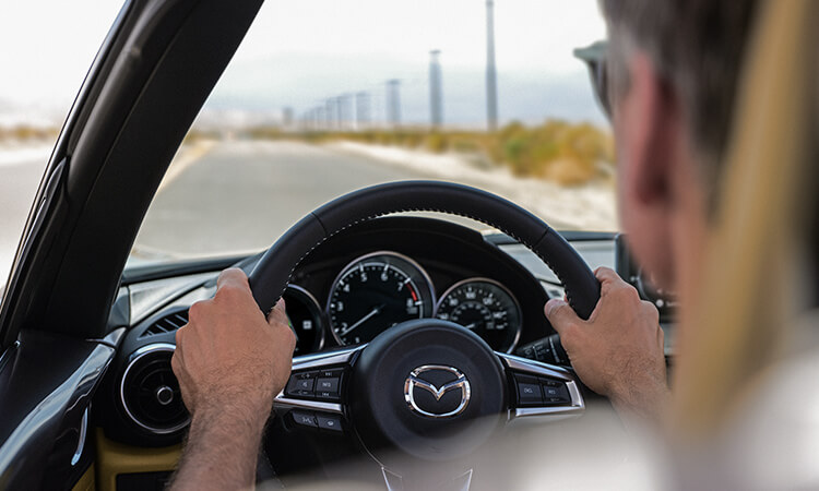Over-the-shoulder view of a person with both hands on the steering wheel of an MX-5 RF driving down a road. 