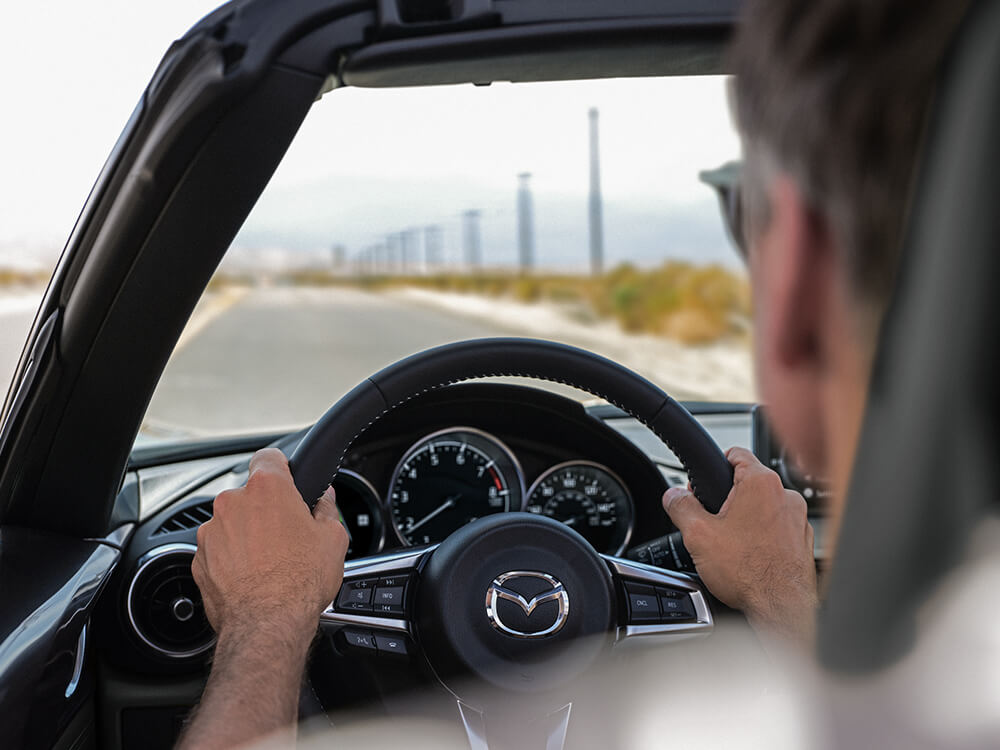 Over-the-shoulder view of a person with both hands on the steering wheel of an MX-5 ST driving down a road.