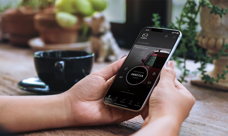 Des mains tenant un téléphone intelligent qui affiche l’écran d’accueil de l’application MyMazda.