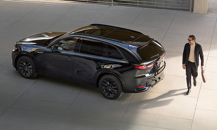 Man with a bag smiles as he walks behind a Jet Black Mica CX-70 parked on daylit concrete parkway.
