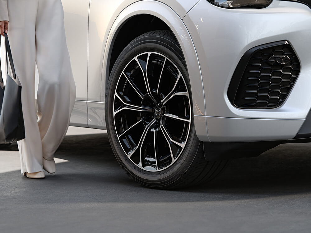 Closeup of front right wheel on a Rhodium White Metallic CX-70, a passenger walks by