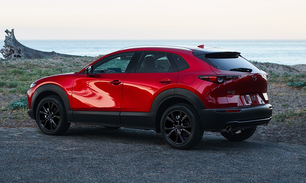 Side view of a Soul Red Crystal Metallic Mazda CX-30 parked near a body of water on a gravel road.