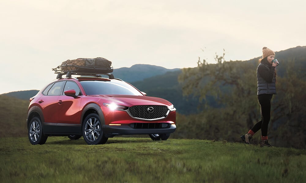 Woman standing next to a Soul Red Crystal Metallic Mazda CX-30 with headlights on, roof rack loaded with gear, in a grassy wildreness setting in the morning.