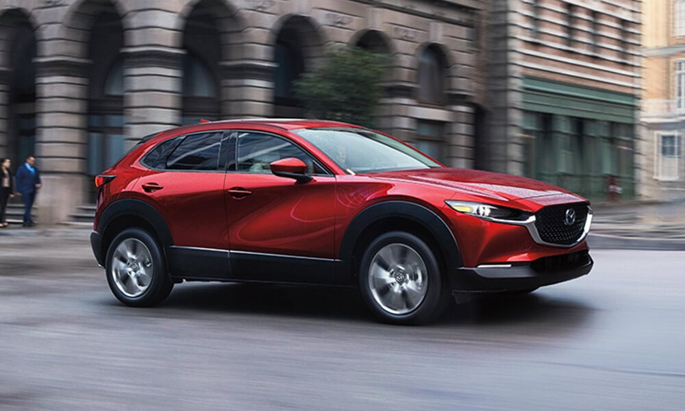 Side profile of a Soul Red Crystal Metallic Mazda CX-30 driving on a city street with buildings and pedestrians blurred to emphasize motion. 