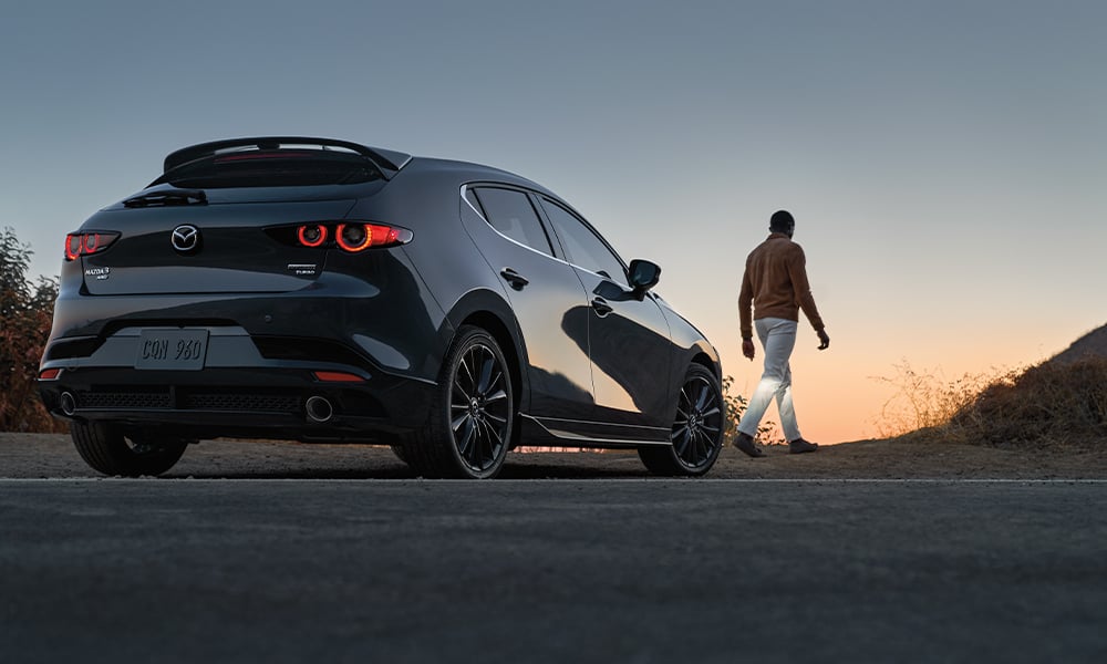 Side profile of a woman wearing sunglasses, seen through the open passenger-side window, driving a Mazda3 Sport.